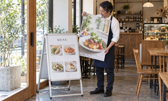 メニュー用スタンド看板の横で交換用ポスターを持つカフェの店主｜誉PRINTING