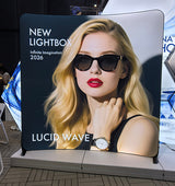 ルーシッド（Lucid）ウェーブの曲線型大型バックパネルにサングラスの女性モデルを配置した展示会での設置例｜誉PRINTING